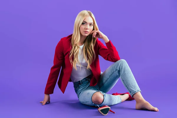 紫色の背景に床に座って赤いジャケットに興味がある若い女性 — ストック写真