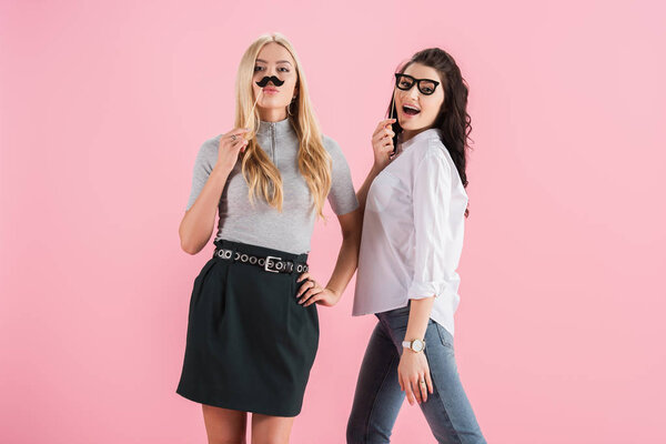 Cheerful girls posing with toy mustache and glasses isolated on pink