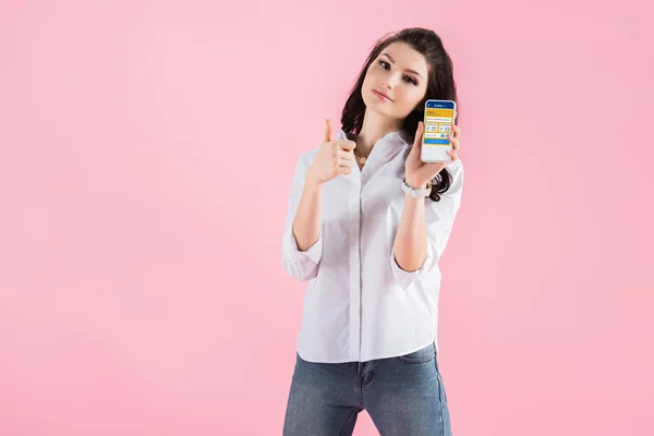 有吸引力的黑发女孩显示拇指和智能手机与预订应用程序在屏幕上 隔离在粉红色 — 图库照片