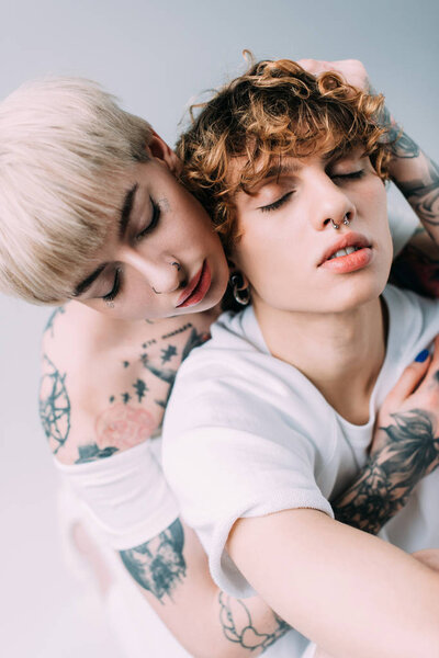blonde woman with tattoos hugging handsome boyfriend with closed eyes isolated on grey
