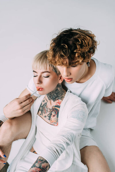 man with curly hair putting cigarette in mouth of blonde tattooed girl isolated on grey