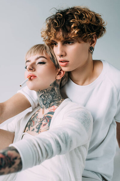blonde tattooed girl holding cigarette in mouth near handsome man with curly hair isolated on grey 