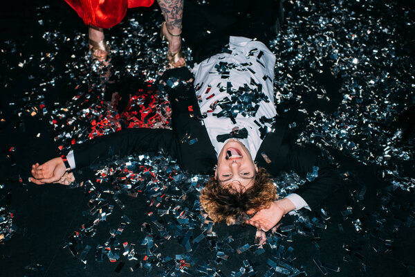 cheerful man lying on floor with confetti and laughing near girlfriend isolated on black