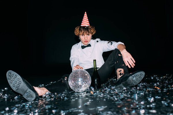 man with curly hair sitting in party cap with disco ball isolated on black