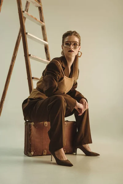 Modische Frau Sitzt Auf Vintage Koffer Neben Leiter Auf Beige — Stockfoto