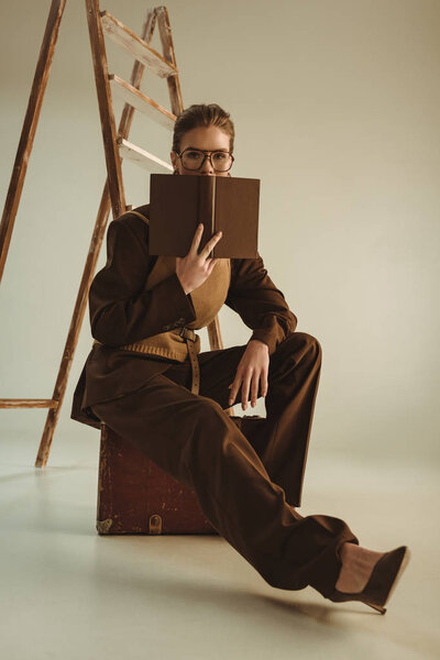 attractive model holding book and sitting near ladder on beige