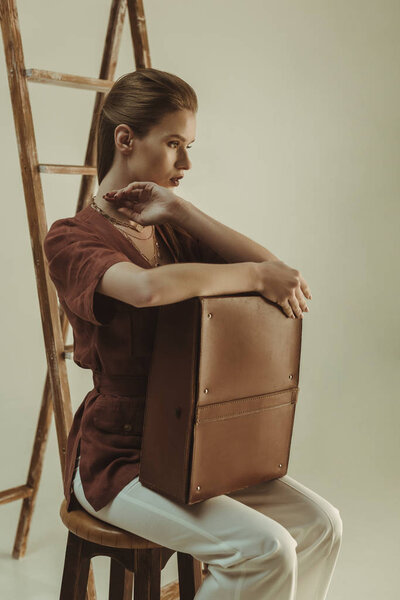 stylish young woman holding leather bag and sitting near ladder isolated on beige