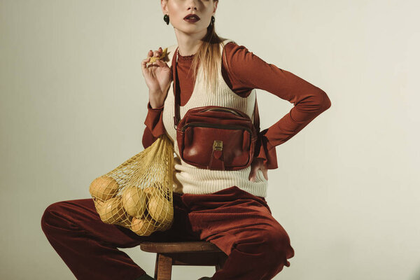 cropped view of fashionable girl sitting on stool with lemons in string bag isolated on beige
