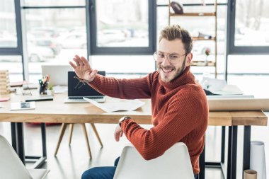 male architect sitting at computer desk, waving and working on blueprints in loft office clipart