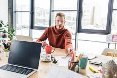 bilgisayar masasında oturan, kameraya bakıyor ve loft Office belge üzerinde çalışan yakışıklı rahat iş adamı