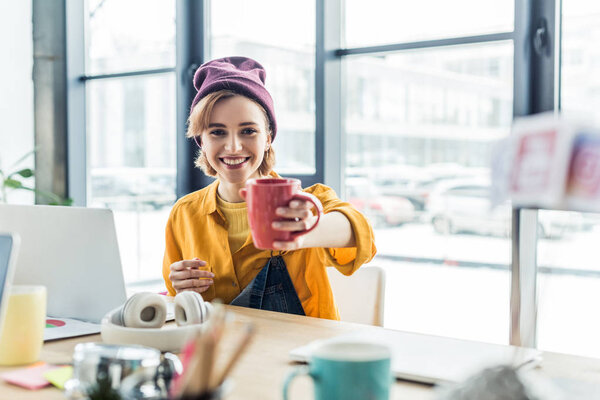 smiling young female it specialist at computer desk holding cup of coffee and looking at camera in loft office 
