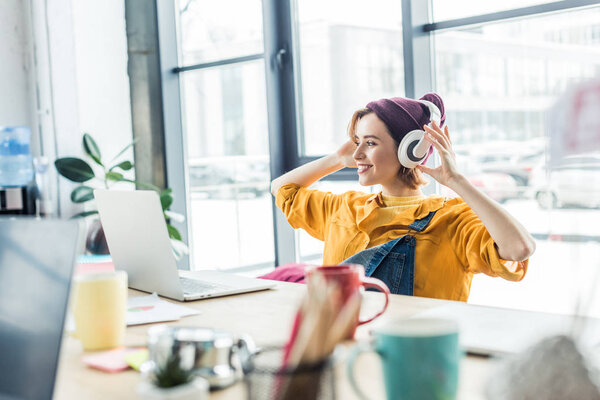young female it specialist in headphones listening music while sitting at computer desk in loft office 