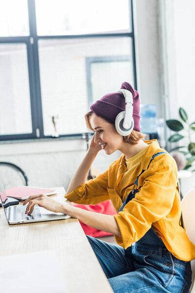 smiling young female it specialist in headphones using laptop in loft office 