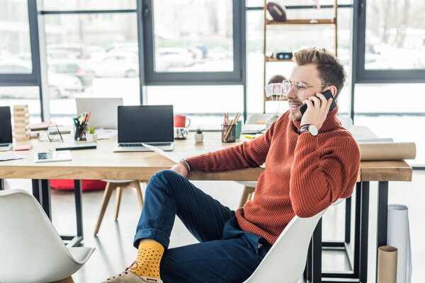 handsome casual businessman sitting at computer desk and talking on smartphone in loft office