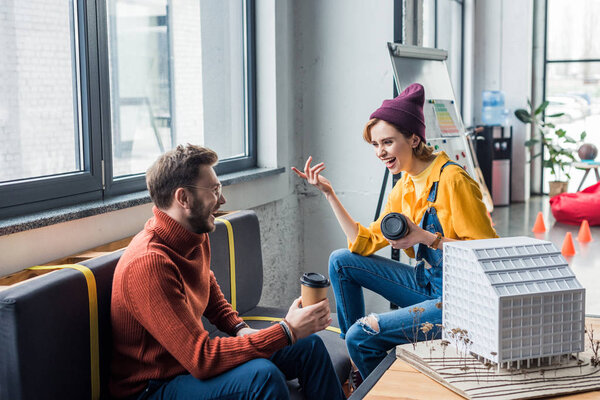 smiling female and male architects with coffee to go working on house model in loft office