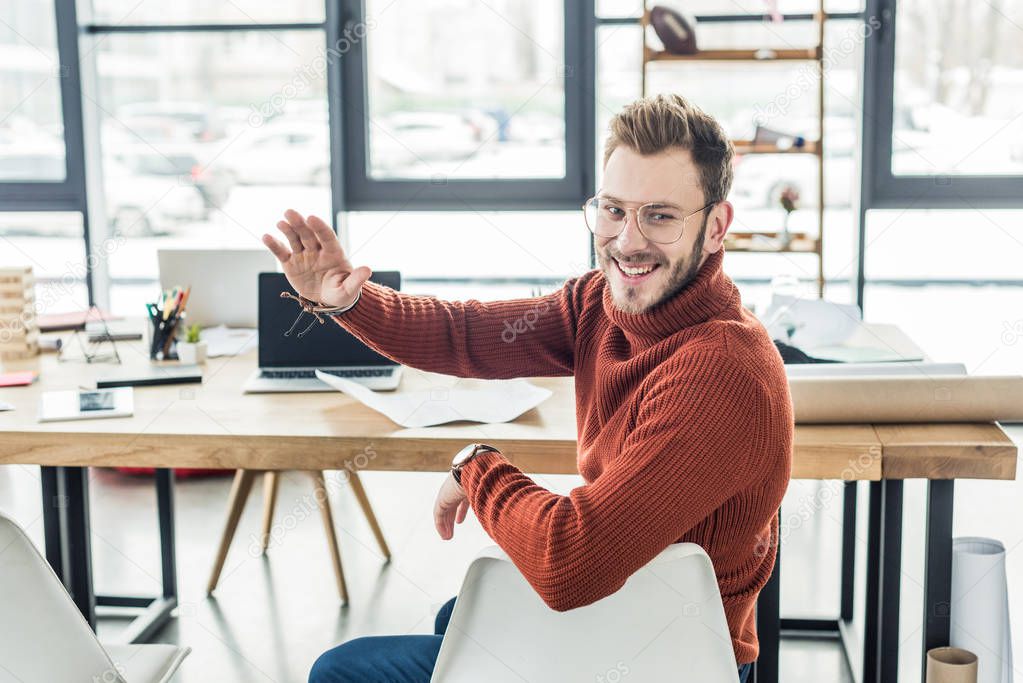 male architect sitting at computer desk, waving and working on blueprints in loft office