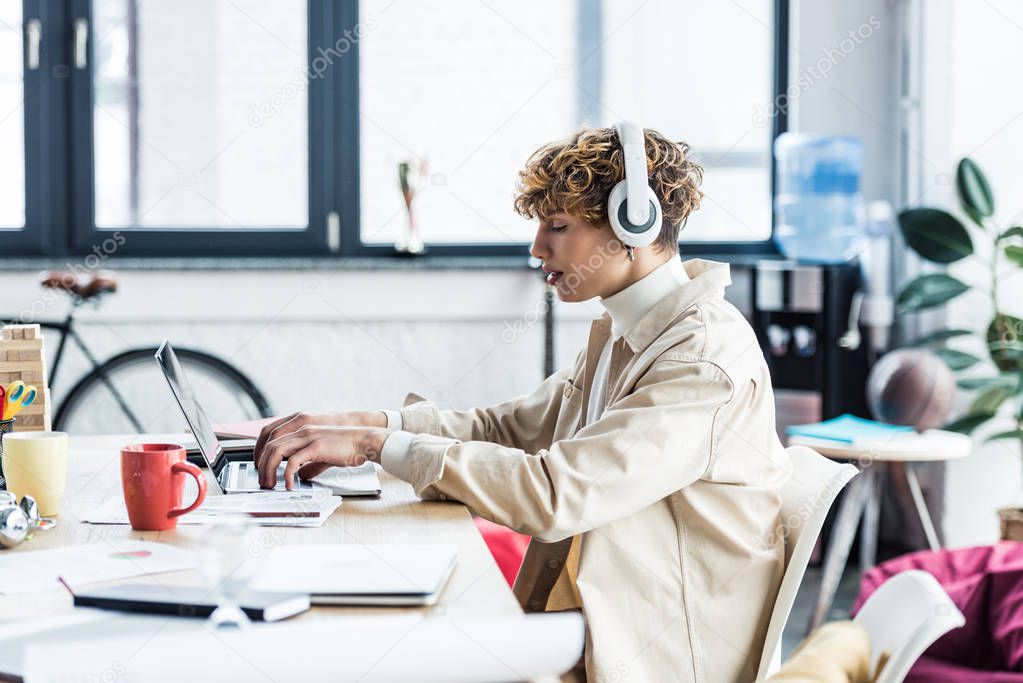 handsome it specialist in headphones sitting at desk and using laptop in loft office