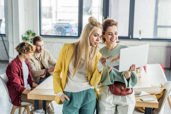 красивые случайные деловые женщины, использующие ноутбук и работающие над проектом запуска в лофт-офисе с коллегами-мужчинами на заднем плане
