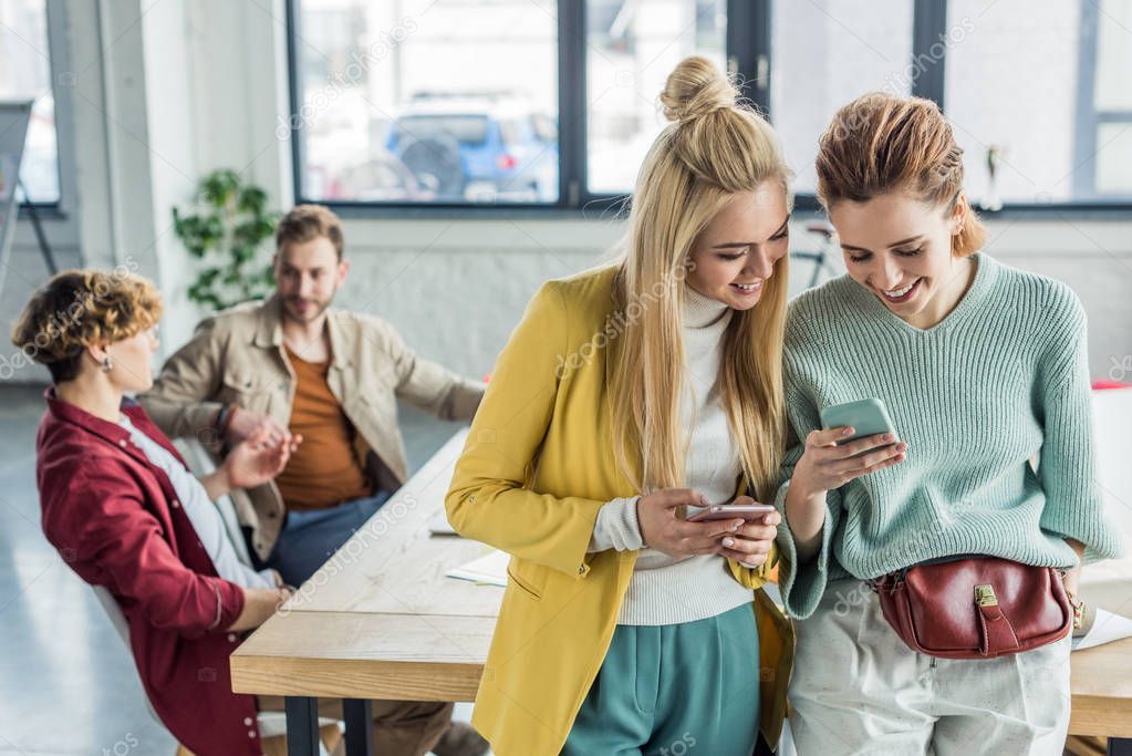 beautiful smiling casual businesswomen using smartphones in loft office with colleagues on background