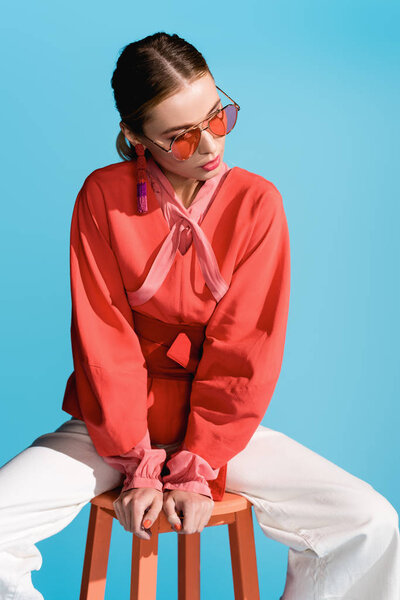 stylish woman in trendy living coral clothing and sunglasses posing on stool isolated on blue