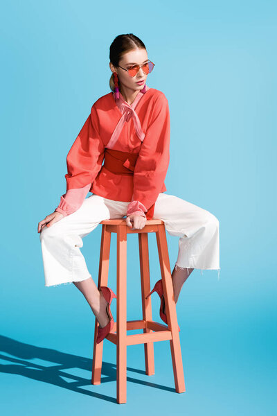 fashionable woman in living coral clothing and sunglasses posing on stool on blue