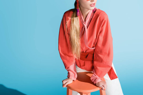 cropped view of fashionable woman in living coral clothing posing on stool isolated on blue