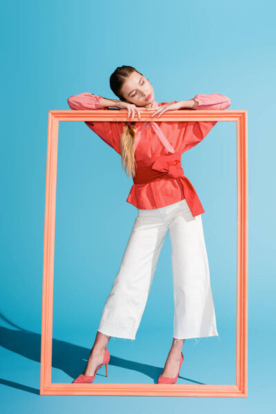 fashionable elegant woman in living coral clothing posing with big frame on blue