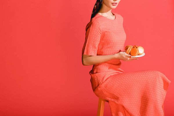 cropped view of stylish girl holding plate with macaroons isolated on living coral. Pantone color of the year 2019 concept