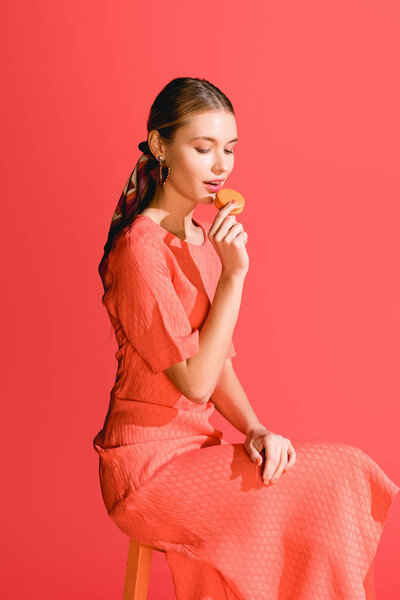 fashionable woman eating macaroon isolated on living coral background