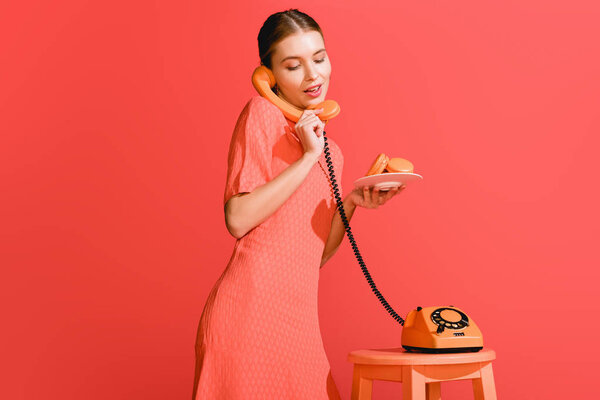 красивая женщина со сладкими макаронами и роторным телефоном изолированы на живых кораллов. Цвет пантона в концепции 2019 года
