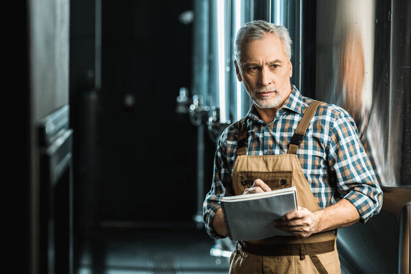 professional senior brewer in working overalls writing in notepad in brewery