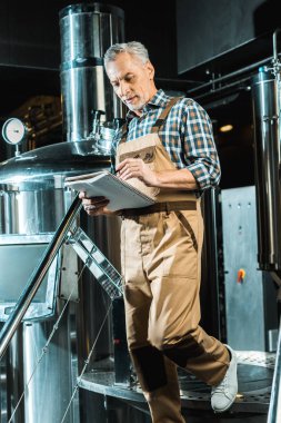 handsome brewer in working overalls writing in notepad while examining brewery clipart