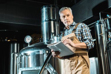 senior brewer in working overalls writing in notepad while examining brewery equipment clipart