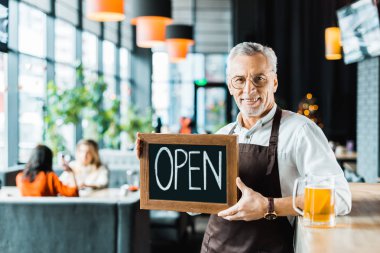 smiling owner of pub holding open sign and standing near bar counter with glass of beer clipart