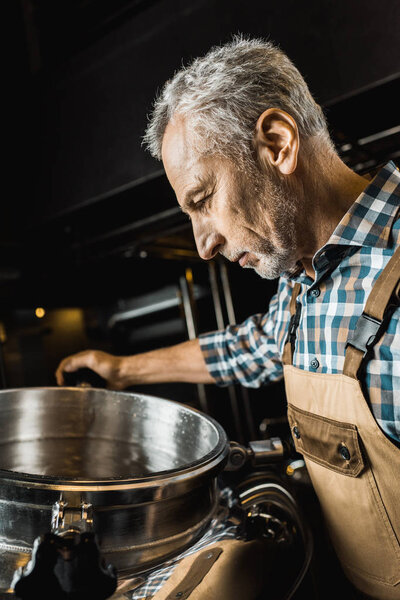 handsome male brewer in working overalls checking brewery equipment