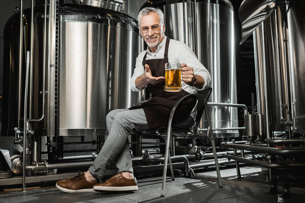 smiling senior brewer in apron sitting on chair and showing glass of beer in brewery