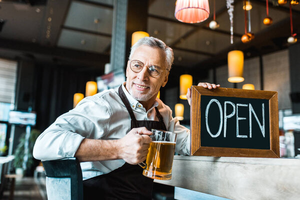senior brewer holding open sign and glass of beer in pub 