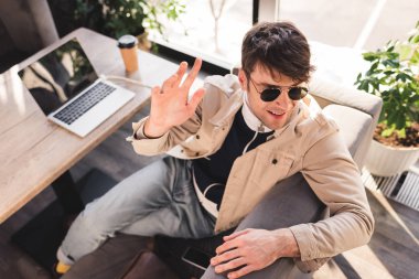 selective focus of trendy man in sunglasses sitting near laptop and paper cup while waving hand in cafe clipart