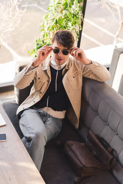 stylish man touching sunglasses while sitting in cafe