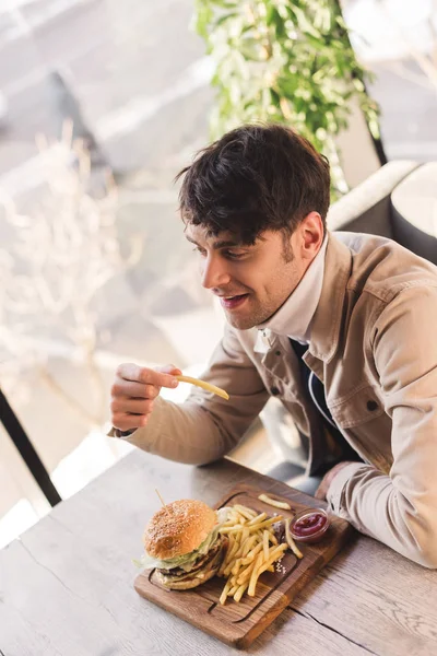 Wesoły Człowiek Posiadający Francuski Fry Pobliżu Smaczne Burger Deski Krojenia — Zdjęcie stockowe