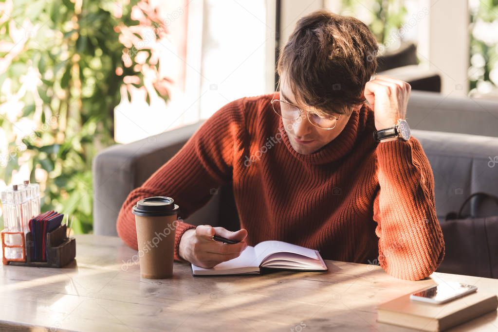 handsome man in glasses looking at notebook near disposable cup in cafe