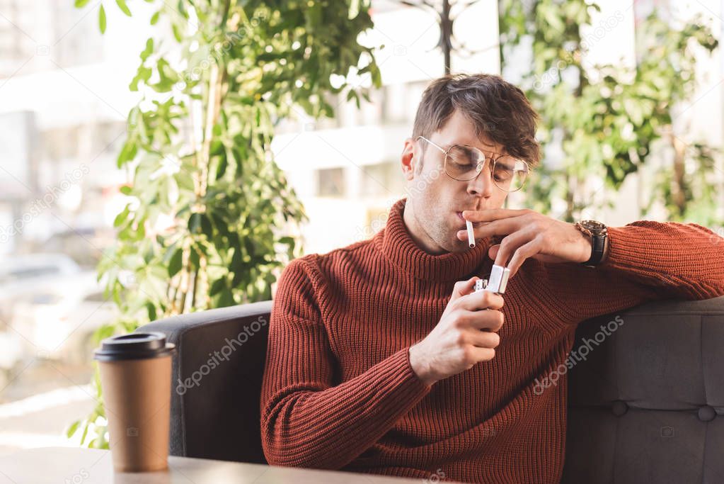 handsome man in glasses smoking cigarette near paper cup with coffee in cafe