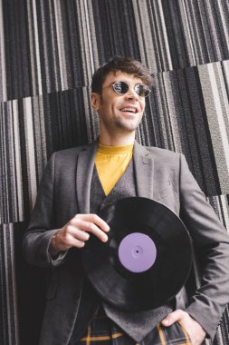 neşeli genç adam plastik retro vinil kayıt tutarak ve elle cebinde duran güneş gözlüğü