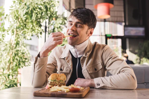 Cheerful Young Man Eating French Fry Tasty Burger Cutting Board — Stock Photo, Image