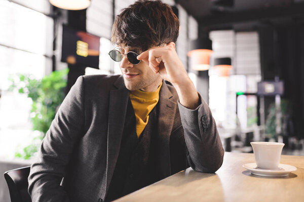 handsome man touching sunglasses and smiling near cup with coffee while sitting at bar counter