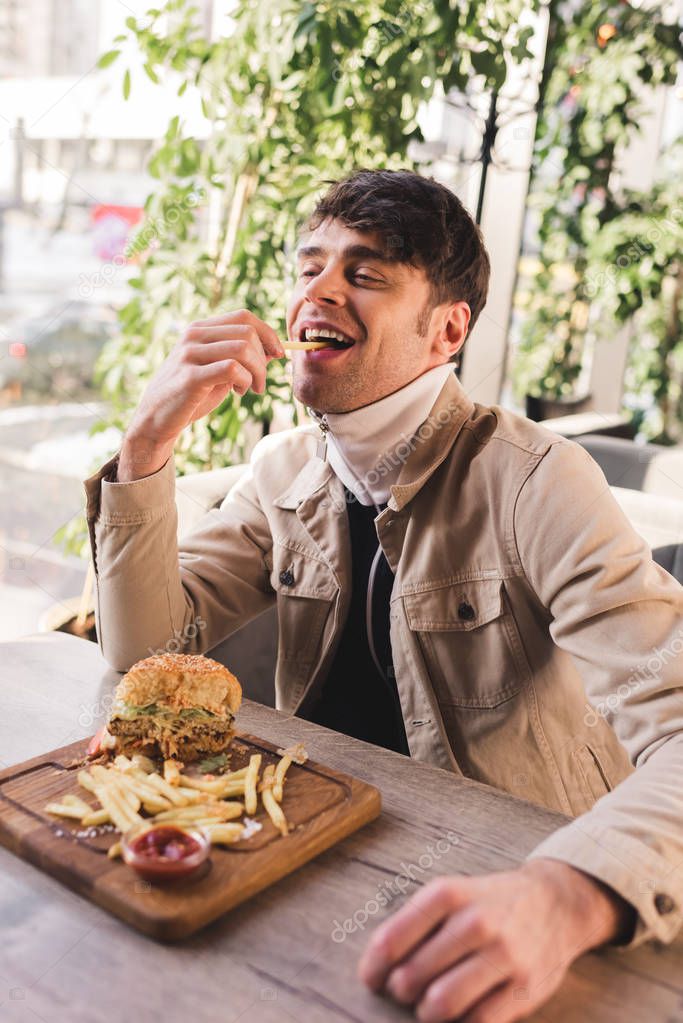 happy man eating french fry near delicious burger on cutting board in cafe
