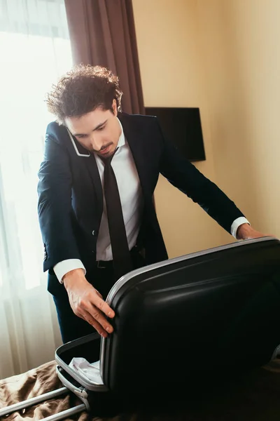 スーツケースはホテルの部屋でベッドの上でスマート フォンに話しているハンサムな実業家 — ストック写真