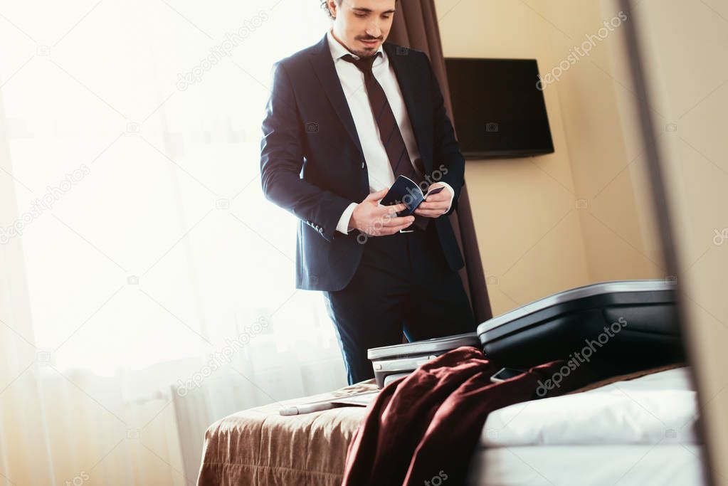 adult businessman holding passport in hotel room with suitcase on bed
