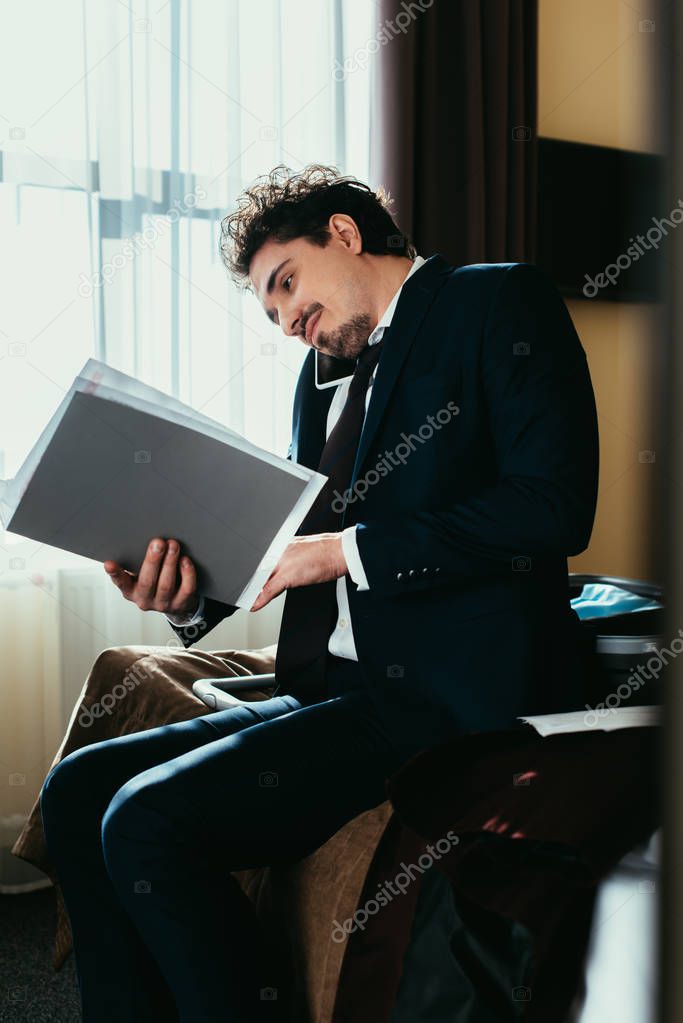 businessman talking on smartphone and holding folder with documents on bed in hotel room