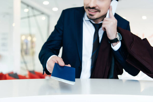 обрезанный вид бизнесмена в костюме с паспортом разговаривая на смартфоне во время регистрации в отеле
